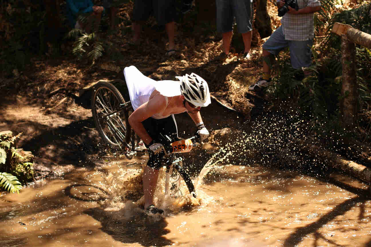 Front, right side view of a cyclist, wearing a dress and riding a Surly bike, straight into a large mud puddle